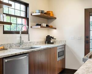 Quartered walnut custom cabinets bring rich wood tones to a kitchen that is otherwise finished with lighter colors and spare finishes, with shelving over the sink